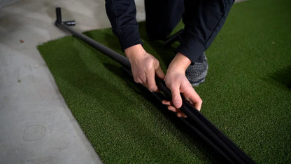 Person assembling black golf simulator frame on artificial turf mat for indoor practice setup