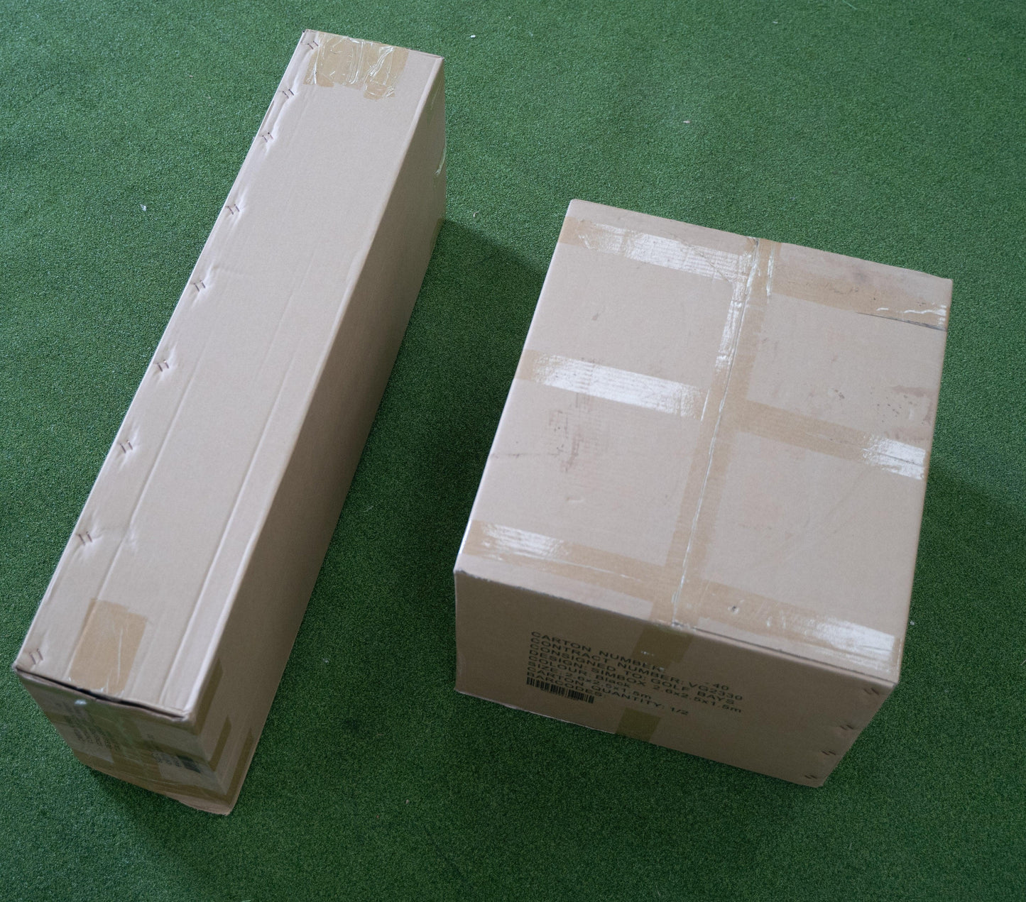 Two packaged golf simulator enclosures on green turf, ready for indoor golf setup