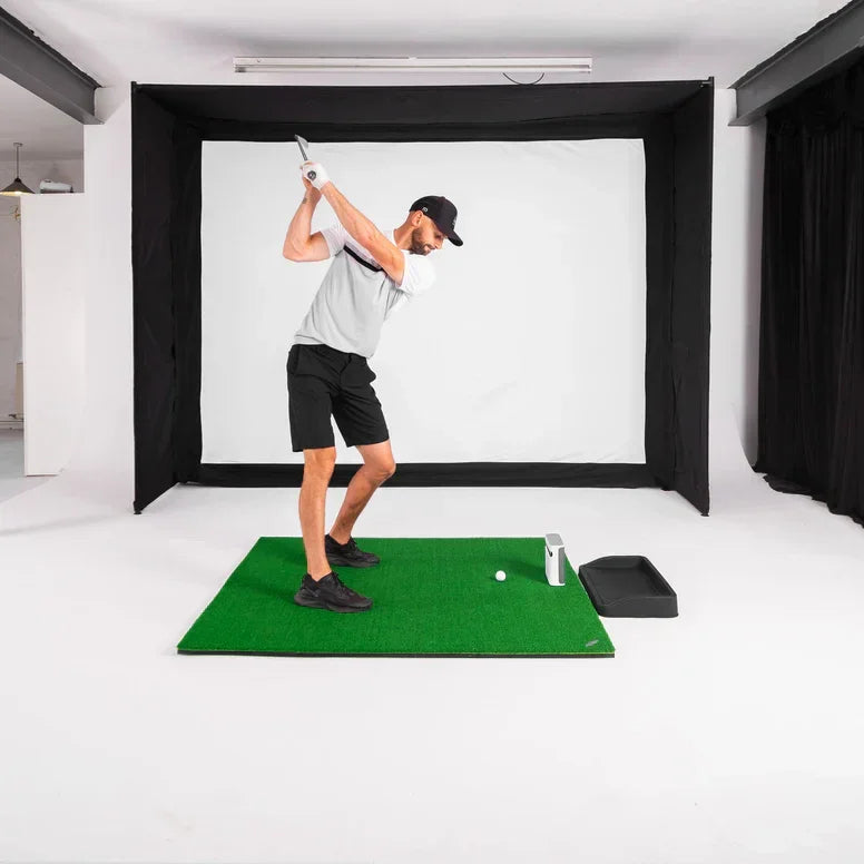 Man swinging golf club on indoor simulator mat with impact screen and golf accessories