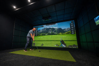 Man practicing golf indoors with simulator, impact screen, and mat in a soundproofed room