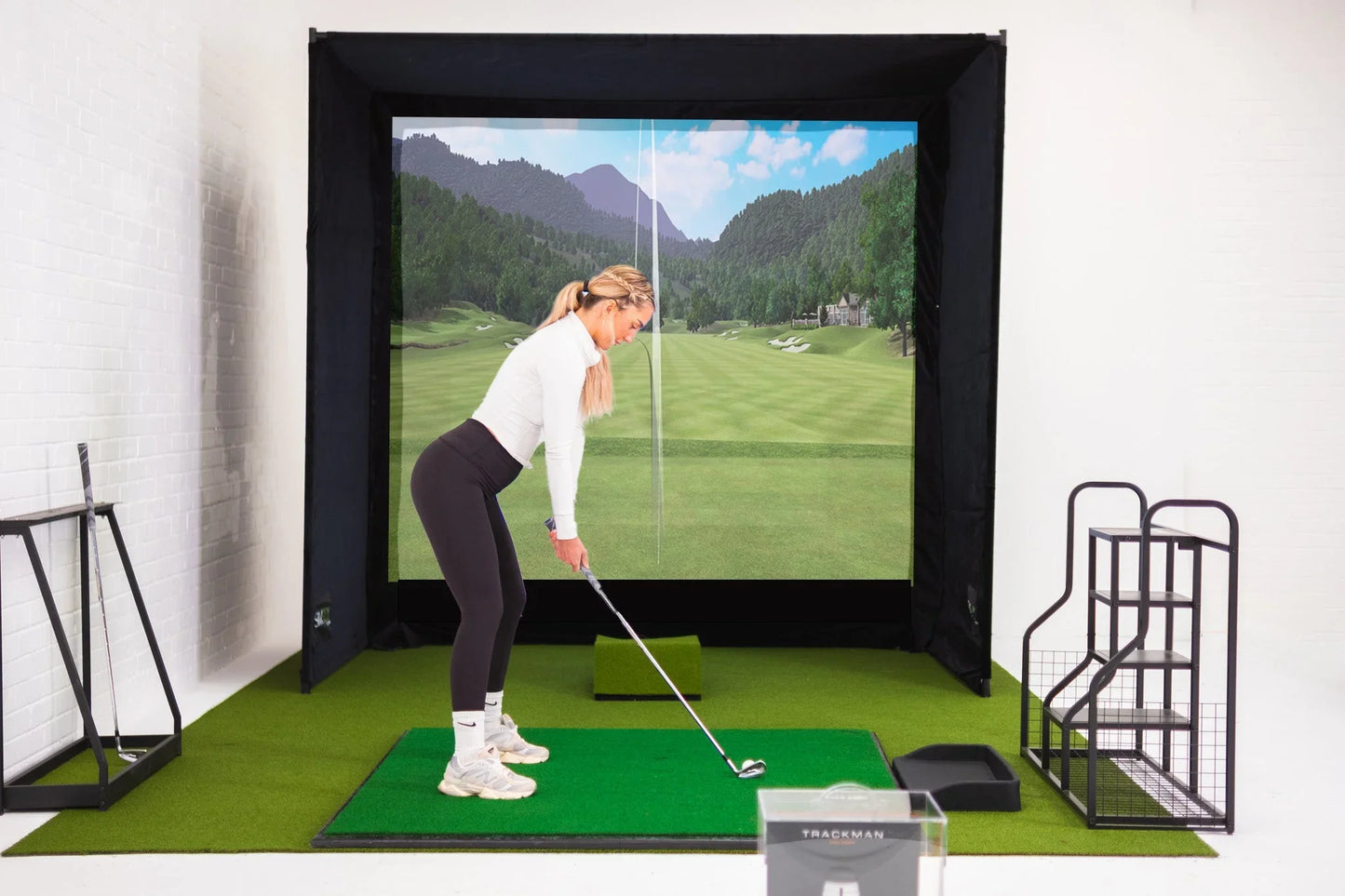 Woman practicing golf swing on indoor simulator with impact screen, mat, and TrackMan device